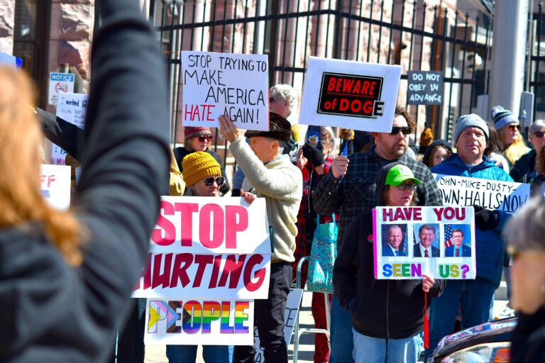 Crowd at a protest holding various signs with political messages in daylight.