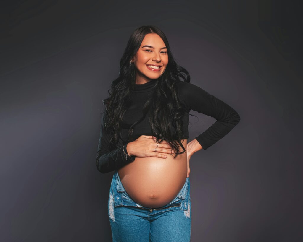 Confident pregnant woman posing and smiling against gray studio background.