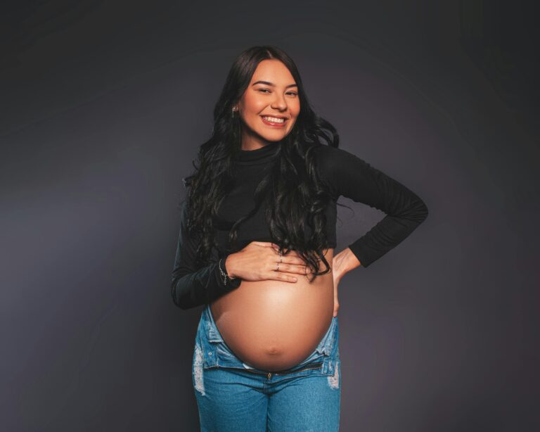 Confident pregnant woman posing and smiling against gray studio background.