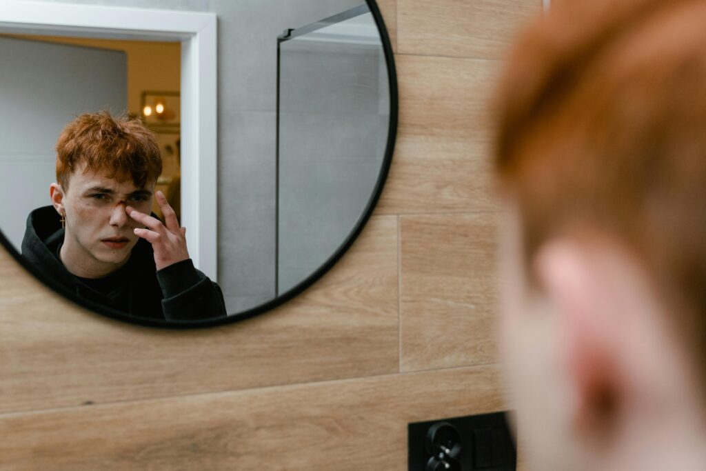 A teenage boy with red hair gazes thoughtfully into a circular mirror indoors.