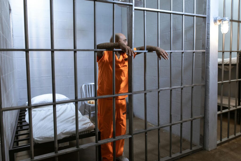 Prisoner in orange uniform standing in a jail cell with iron bars.