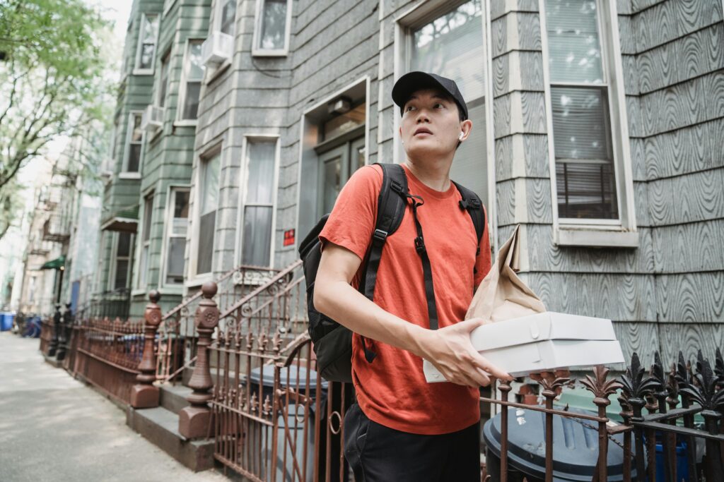 A delivery worker in a red shirt and cap holding packages outside a residential building.