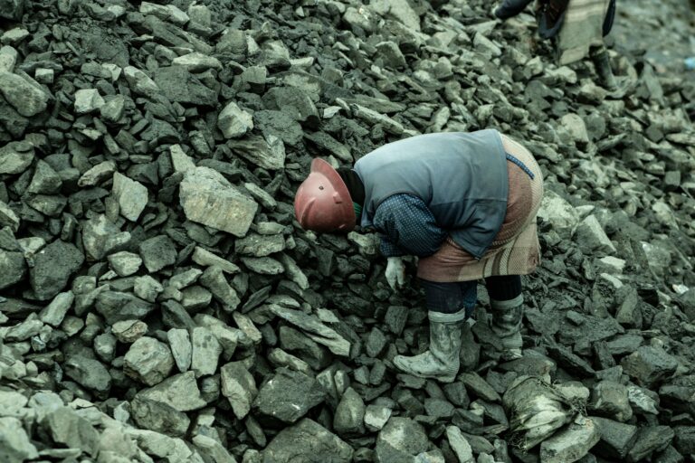 An industrial worker wearing a helmet bends over a heap of stones in a quarry setting.