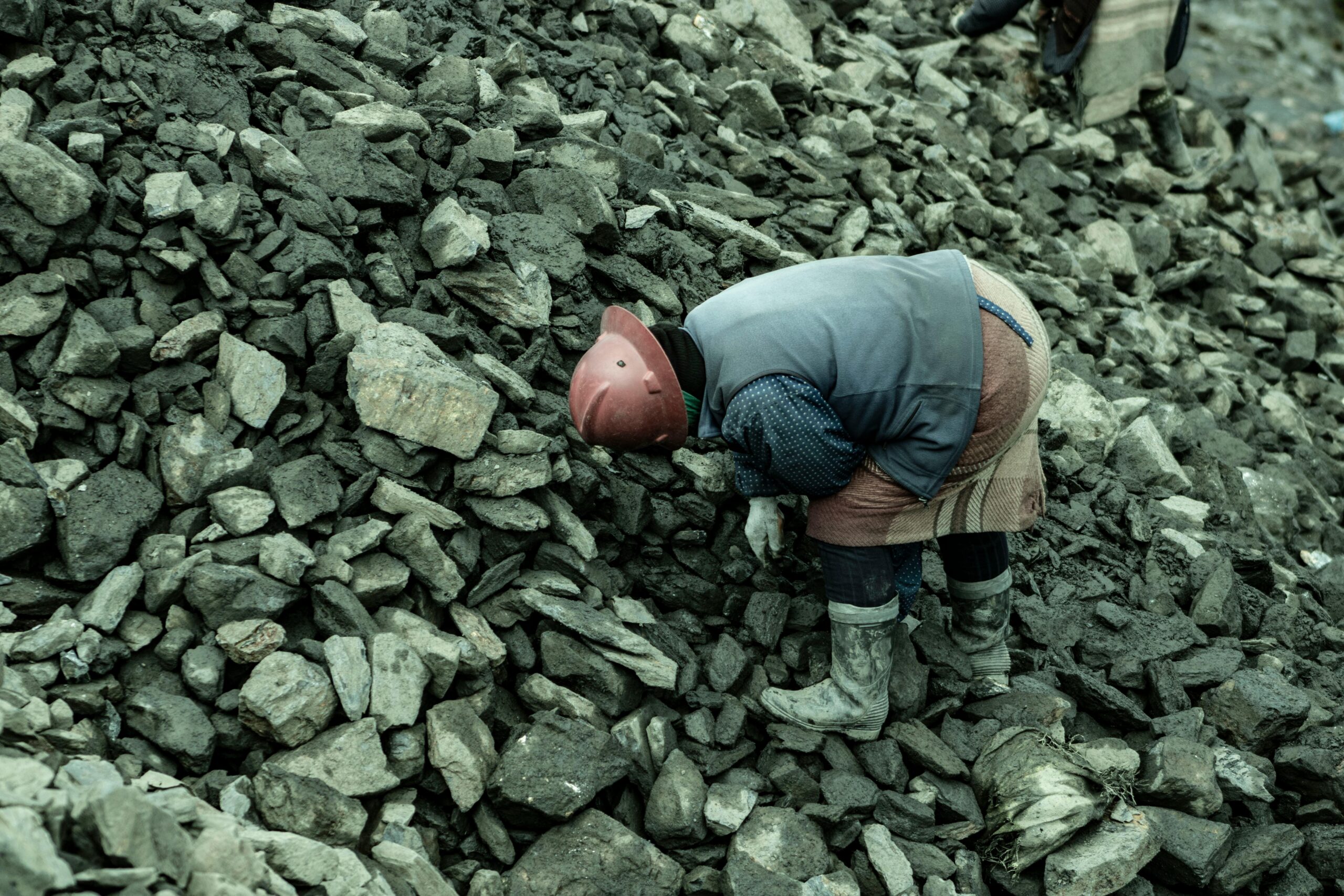 An industrial worker wearing a helmet bends over a heap of stones in a quarry setting.