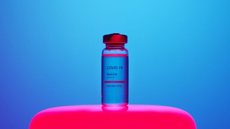 A COVID-19 vaccine vial sits on a bright, blue and pink background. Modern and impactful.