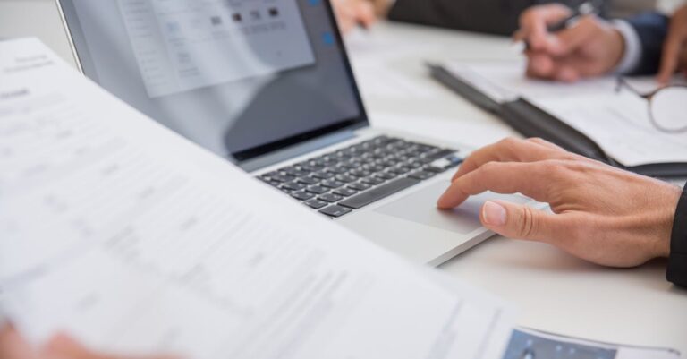 Close-up of hands working on documents and a laptop in an office setting, illustrating teamwork and productivity.